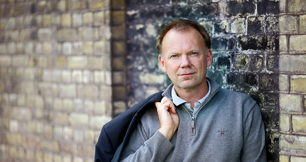 Porträtt Anders Ynnerman står lutad mot en tegelvägg. Han har grå tröja och håller en kavaj över axeln